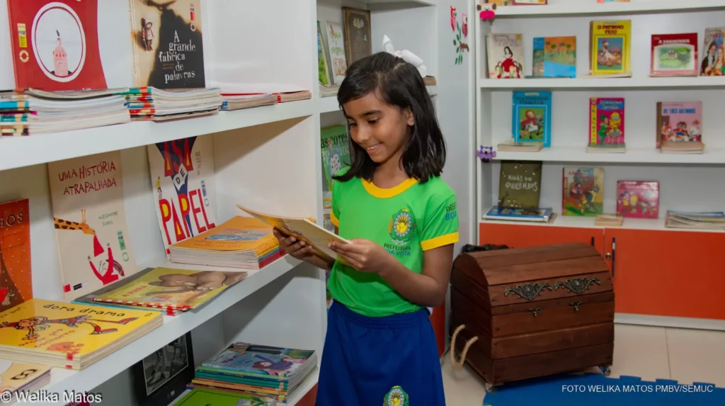 Criança sorridente lendo o livro O Pequeno Príncipe na Biblioteca Municipal de Boa Vista no Tancredo Neves.
